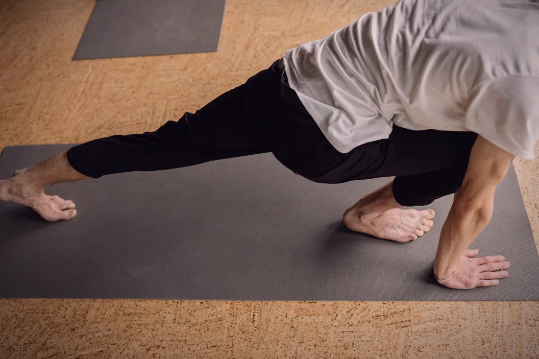A student holds a low lunge on a mat, mid-flow, in a warm-toned studio.