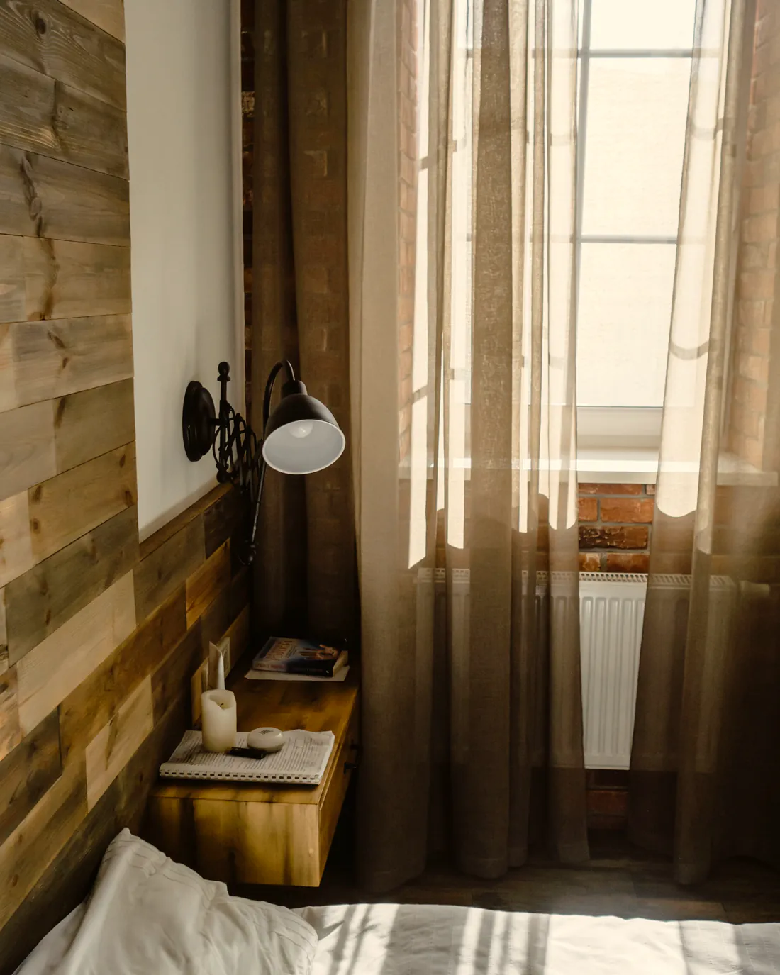 A bedroom corner with white linen, a reclaimed-wood wall and a reading lamp by a sunlit window.