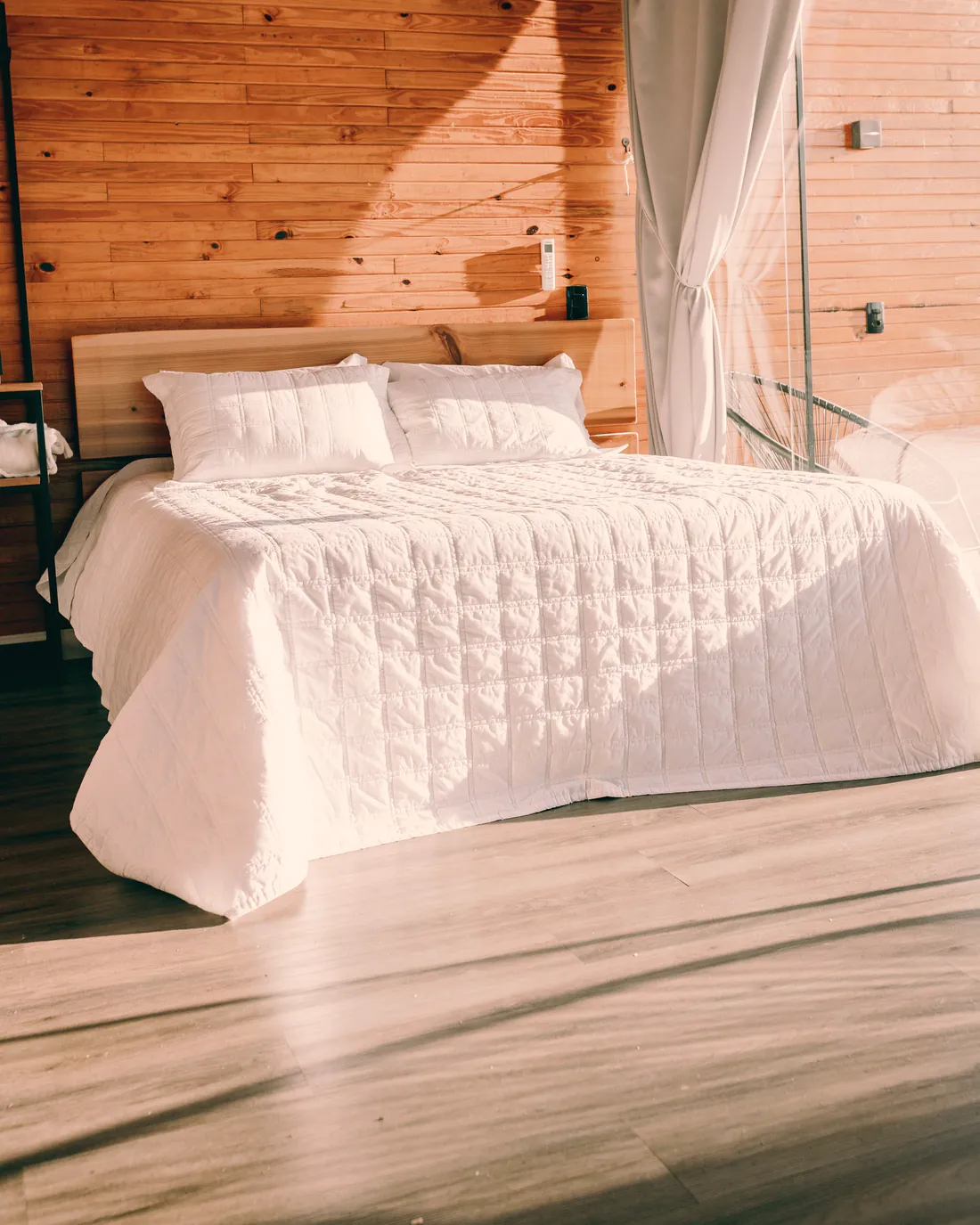 A guest bedroom with crisp white bedding against a pine-clad wall, lit by warm afternoon sun.